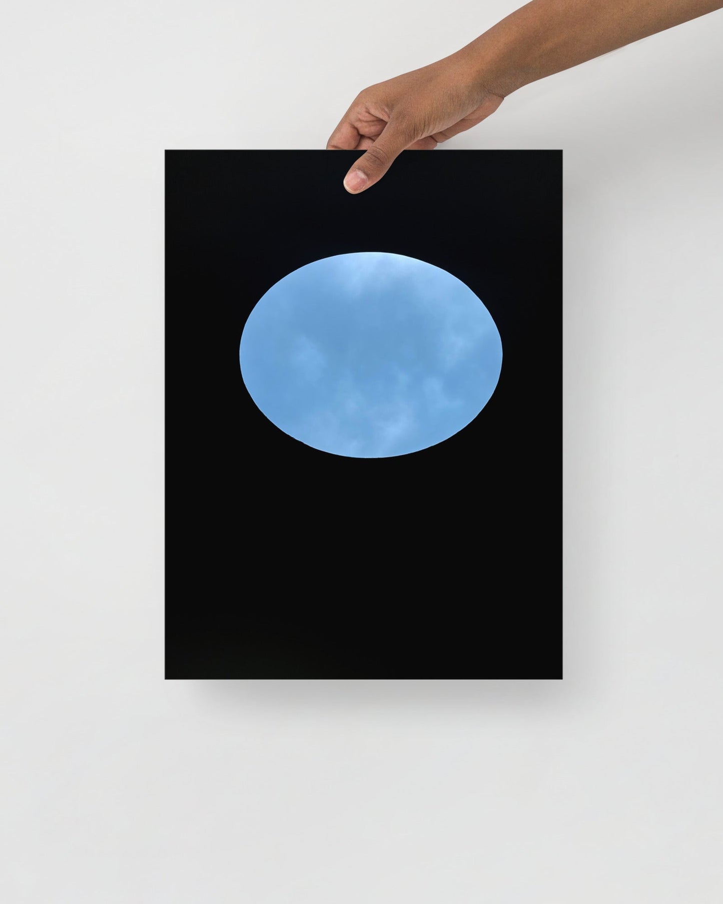 Framed artwork of a blue sky view of oculous in roof of skyspace three gems, held by a hand against a white wall.