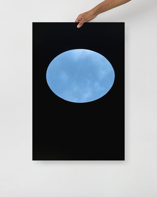 Framed artwork of a blue sky view of oculous in roof of skyspace three gems, held by a hand against a white wall.