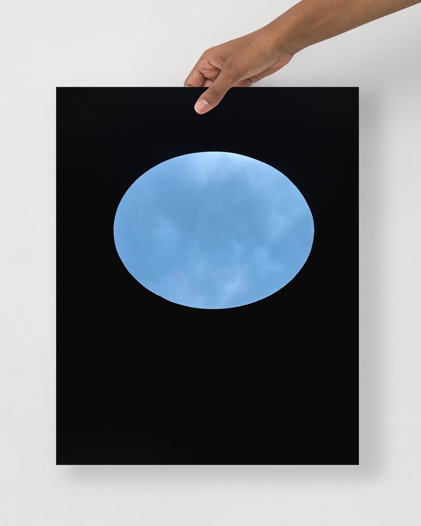 Framed artwork of a blue sky view of oculous in roof of skyspace three gems, held by a hand against a white wall.