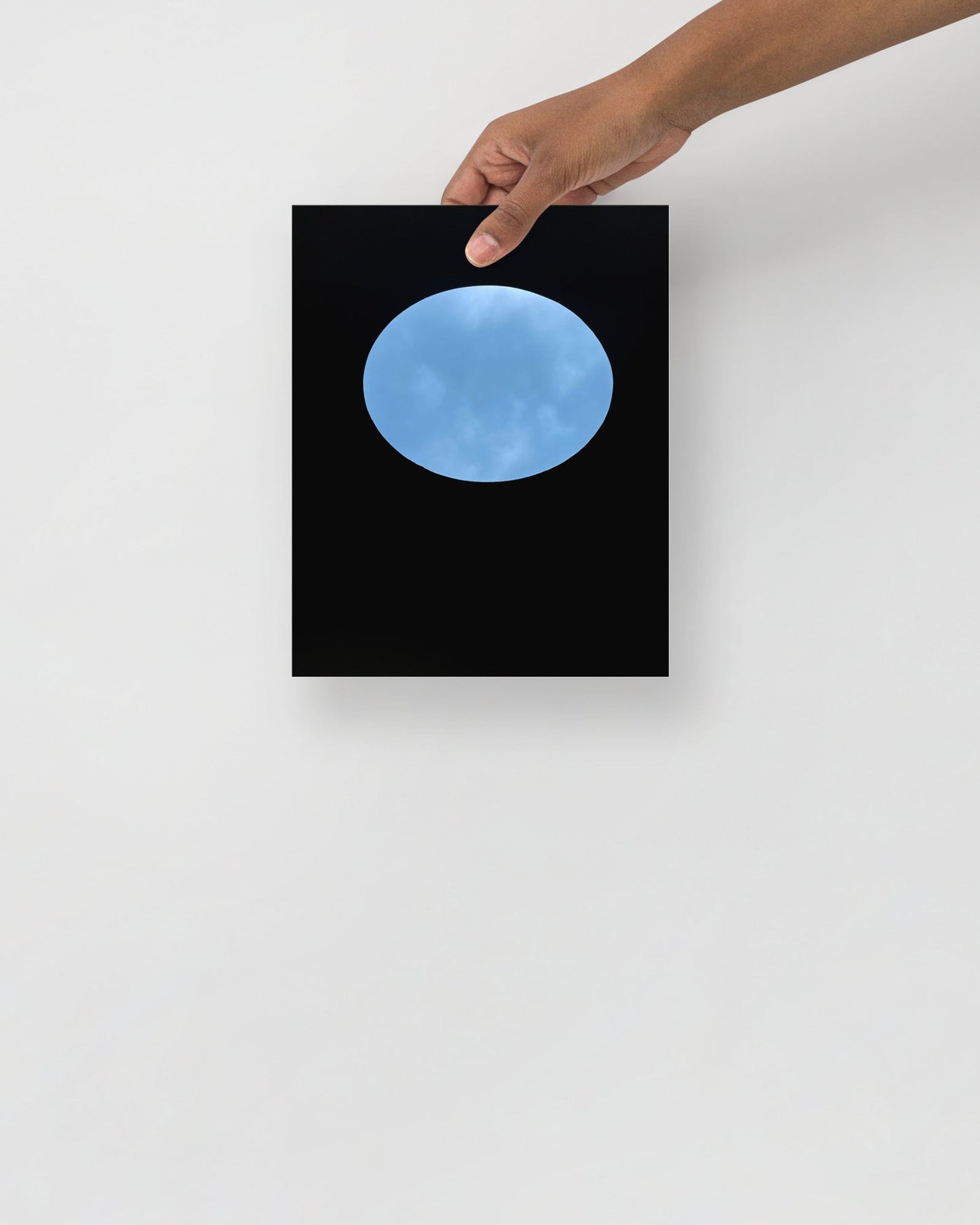 Framed artwork of a blue sky view of oculous in roof of skyspace three gems, held by a hand against a white wall.
